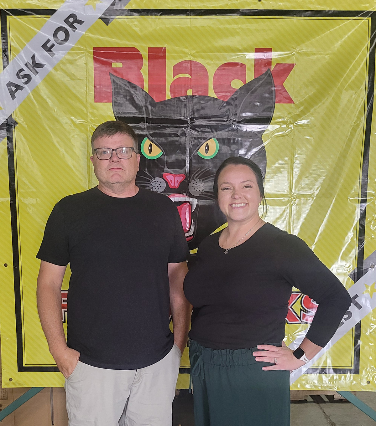 Neil and Meghan of Allstar Fireworks in Missouri standing in front of a yellow banner featuring the BlackCat logo, representing their family-owned fireworks business.